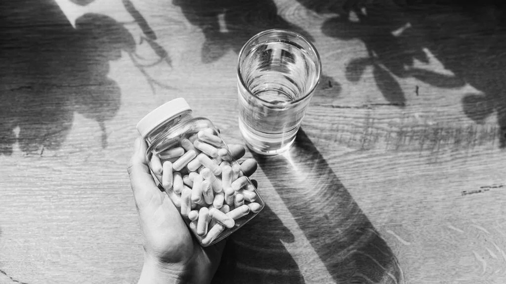 black and white photo of hand holding jar of supplement capsules next to a glass of water